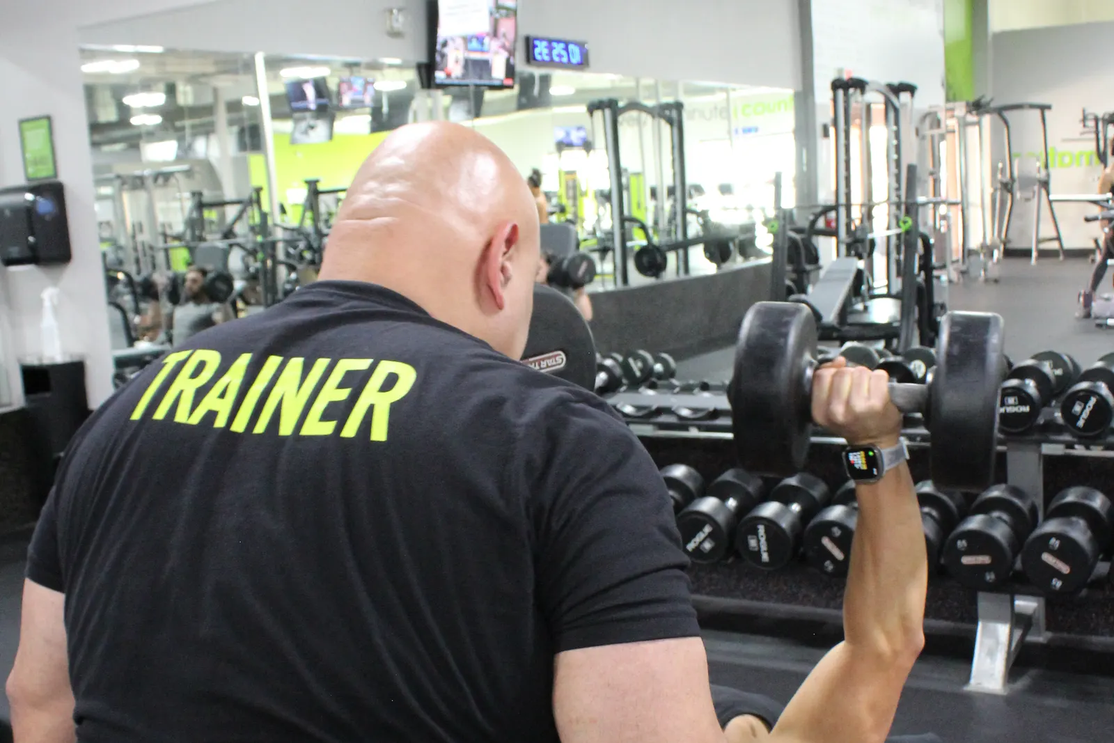 A trainer, seen from behind, spotting someone who is pressing a dumbbell on a bench.