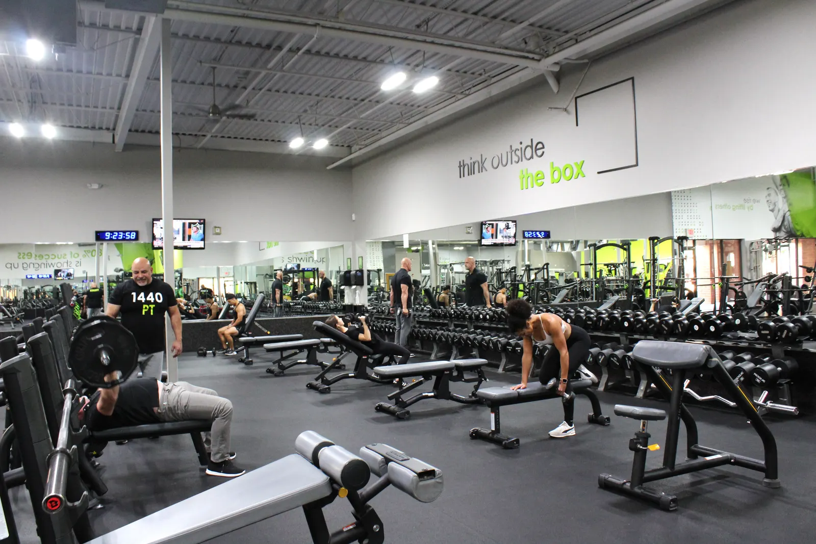 The free-weight floor in use: someone pressing a barbell on a bench, a woman rowing a dumbbell, and dumbbells racked along the mirrored wall.