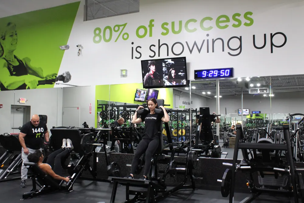 A trainer coaching somebody on the leg press, with the weight machines and the mural behind them.