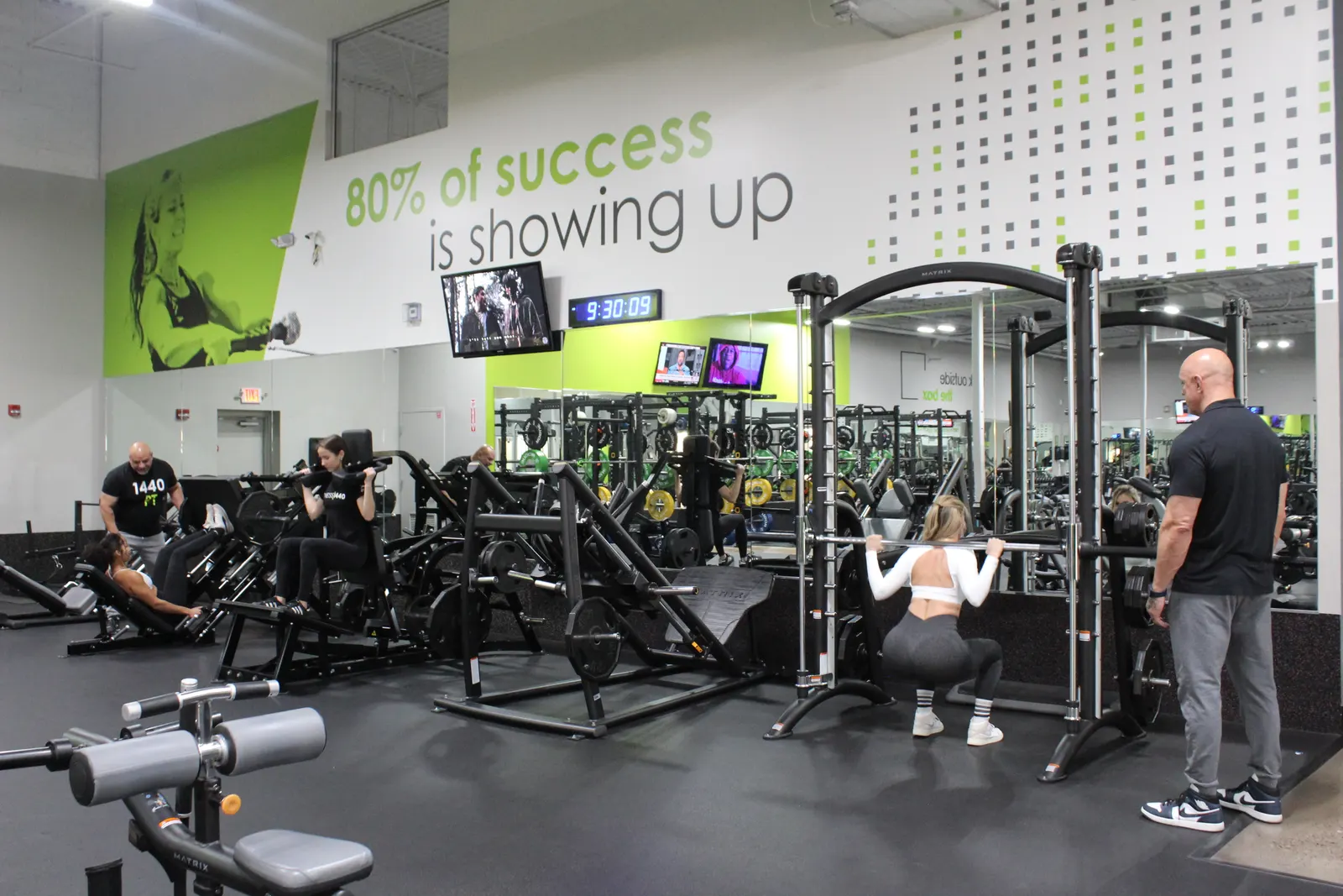 A woman squatting in a rack while a coach watches, with plate-loaded machines in use behind them.