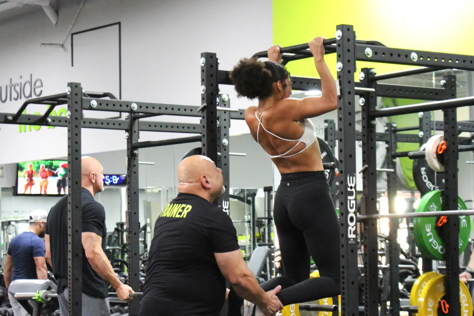 A trainer steadying a woman by the feet as she does a pull-up on a rack.