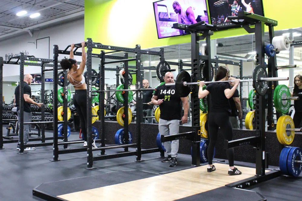 A woman with a loaded barbell across her back at a squat rack, with coloured plates stored on the uprights.