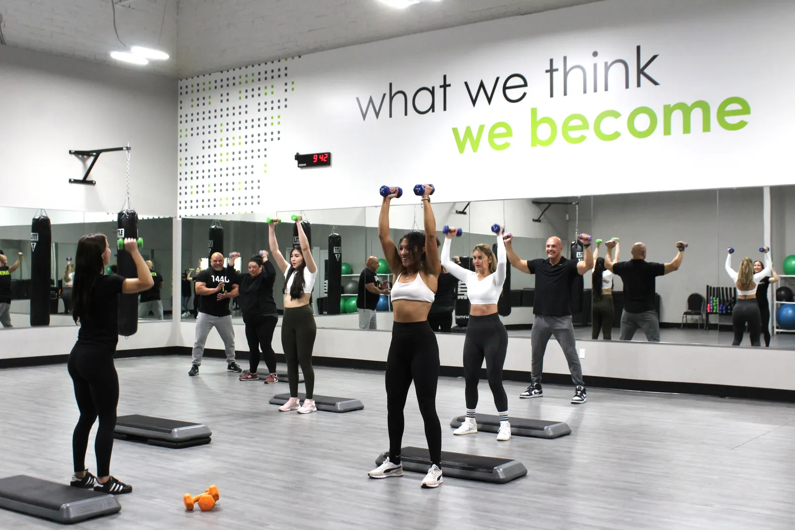 A group in the studio pressing dumbbells overhead, each standing behind a step, with mirrors along the wall.
