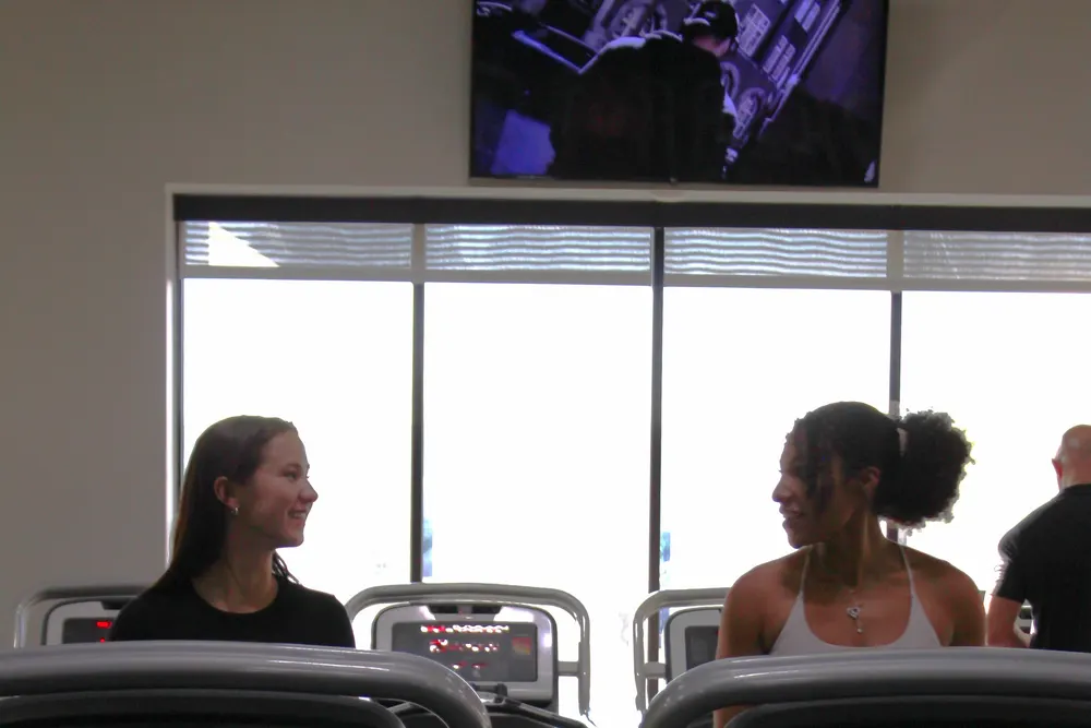 Two women on neighbouring treadmills talking to each other, with a bright window behind them.