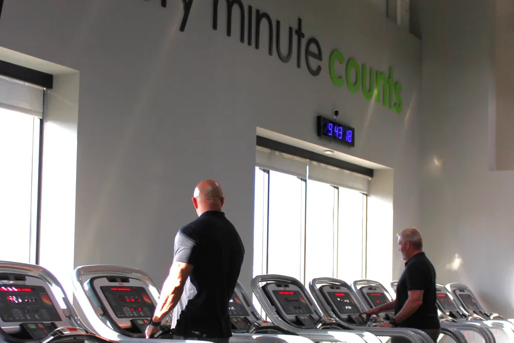 Two men walking on treadmills beside tall windows, seen from behind, with sunlight across the machines.