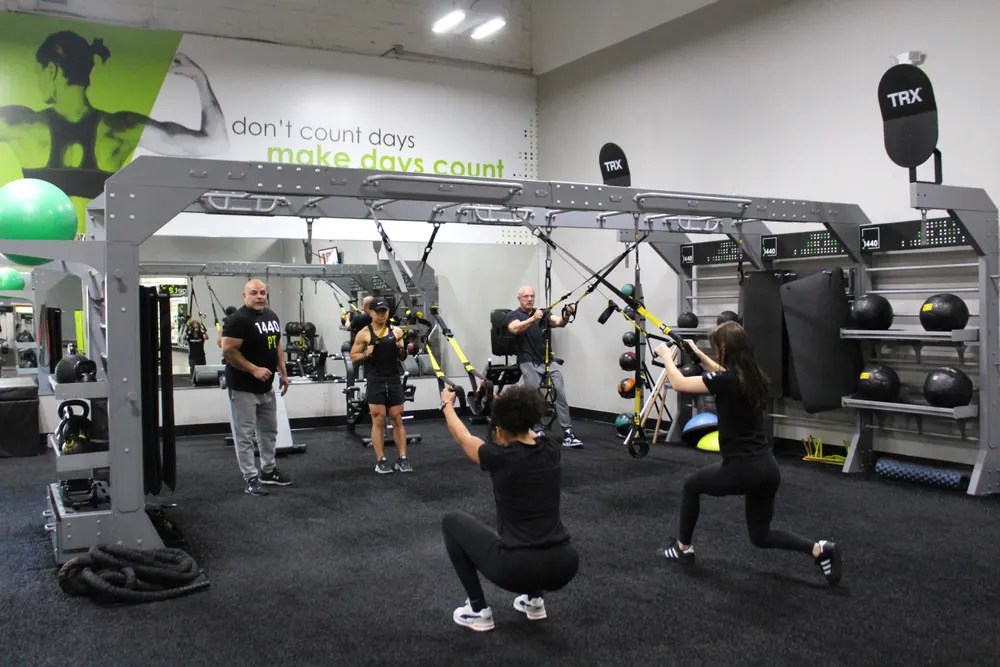 People training on suspension straps at the rig on the turf, one in a deep squat and one in a lunge.
