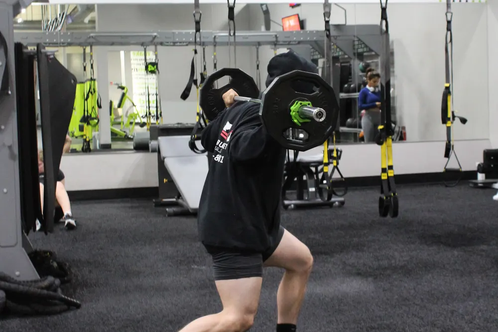 Someone in a hooded top lunging across the turf with a barbell on their back.