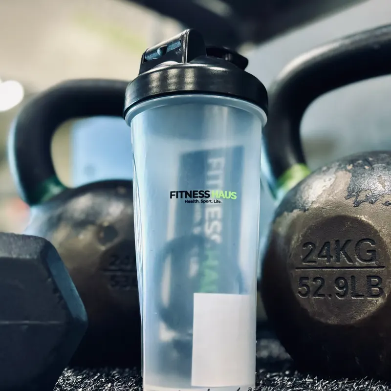 A clear shaker cup with the gym’s logo, on the turf between two kettlebells and a dumbbell.